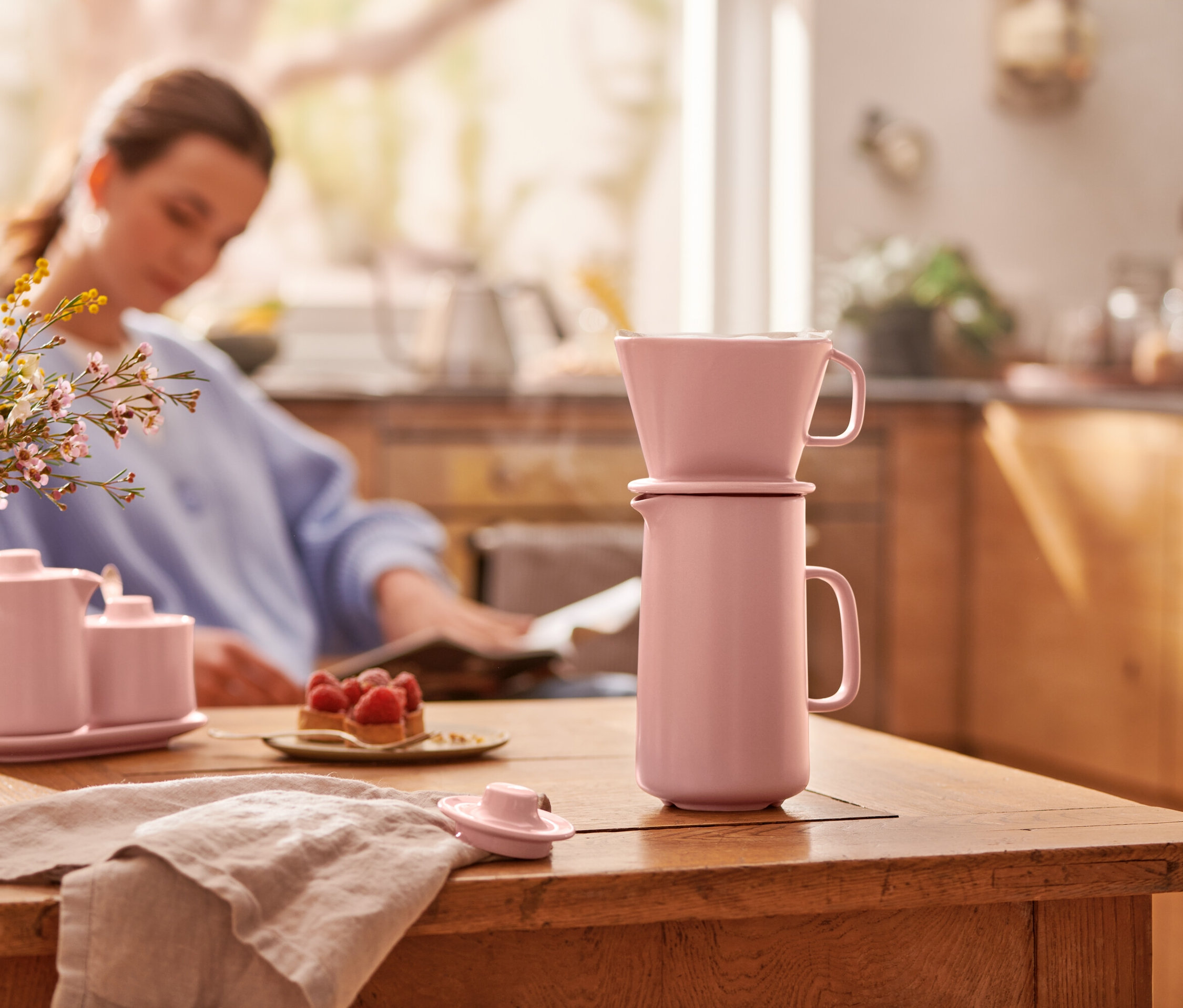 Eine rosa Kaffeekanne mit Filter steht auf einem Holztisch. Im Hintergrund sitzt eine Frau.