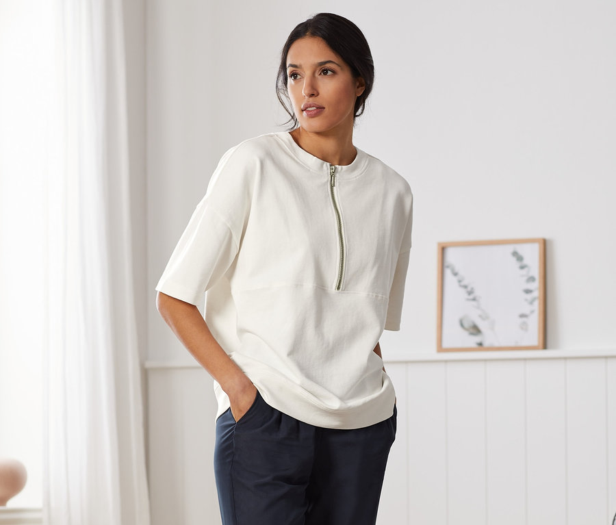 Eine Frau steht in einem Raum und trägt ein weißes Oversized-Sweatshirt, dunkelblaue Hosen und weiße Turnschuhe.