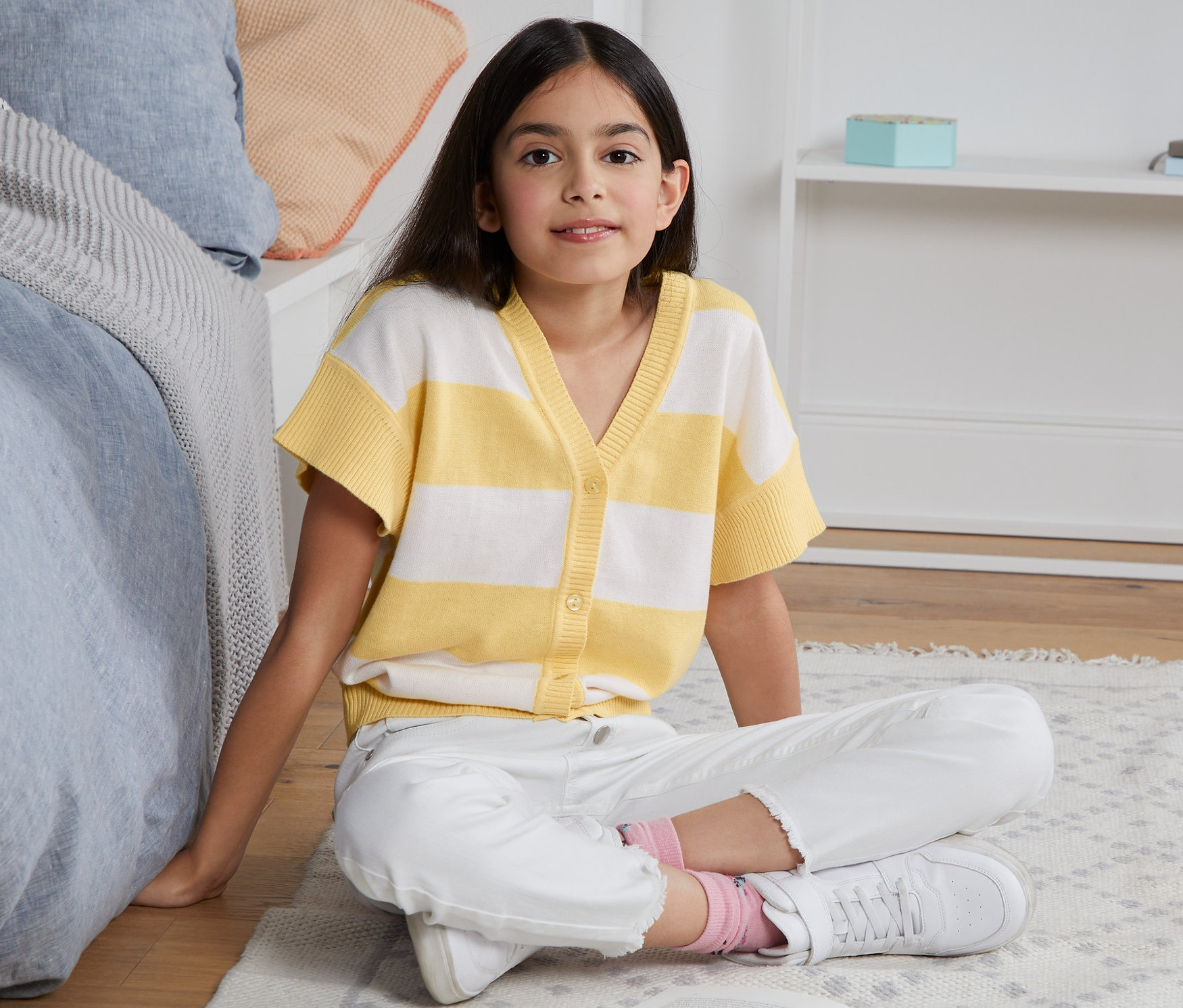 Ein Mädchen sitzt auf dem Boden und liest ein Buch. Sie trägt eine Strickjacke mit kurzem Arm.