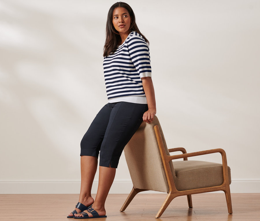 Eine Frau sitzt auf der Lehne eines Stuhls und trägt einen blau-weiß gestreiften Feinstrick-Pullover, eine 3/4-Stretchhose und Sandalen mit Schnalle.