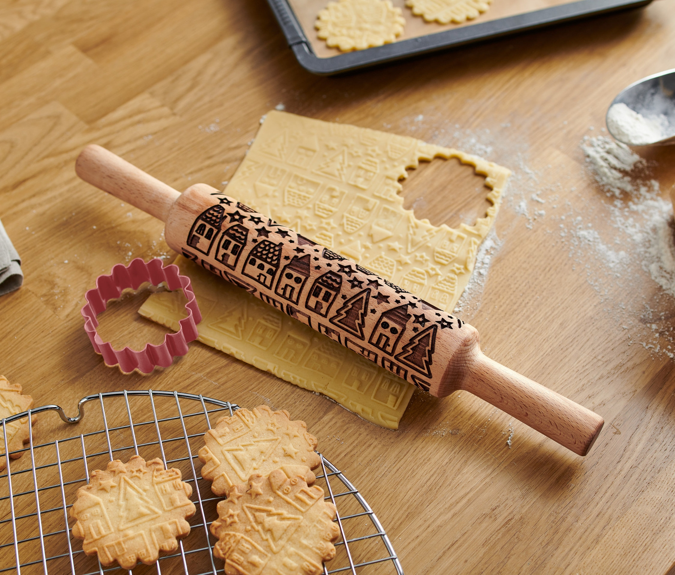 Auf einem Holztisch liegen eine Motiv-Teigrolle, ein Ausstecher und gebackene Kekse.