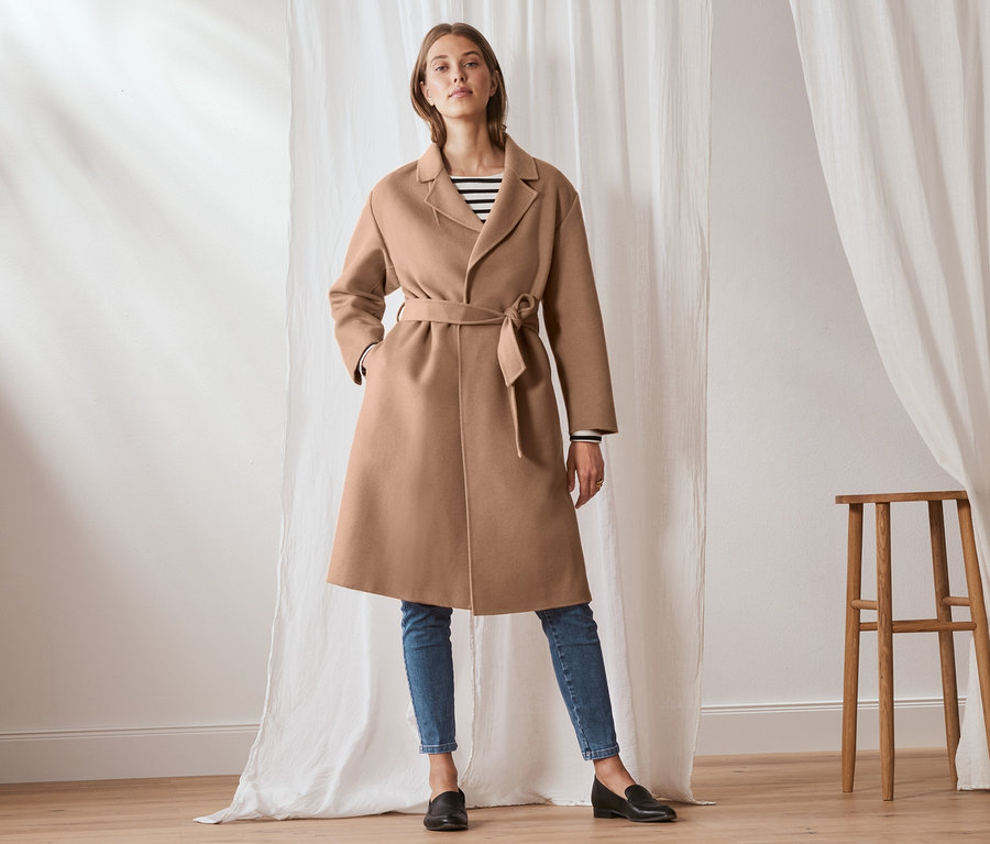 Frau posiert in einem hellbraunen Wollmantel mit Kaschmir und Gürtel, den sie über einem gestreiften T-Shirt und Jeans trägt.