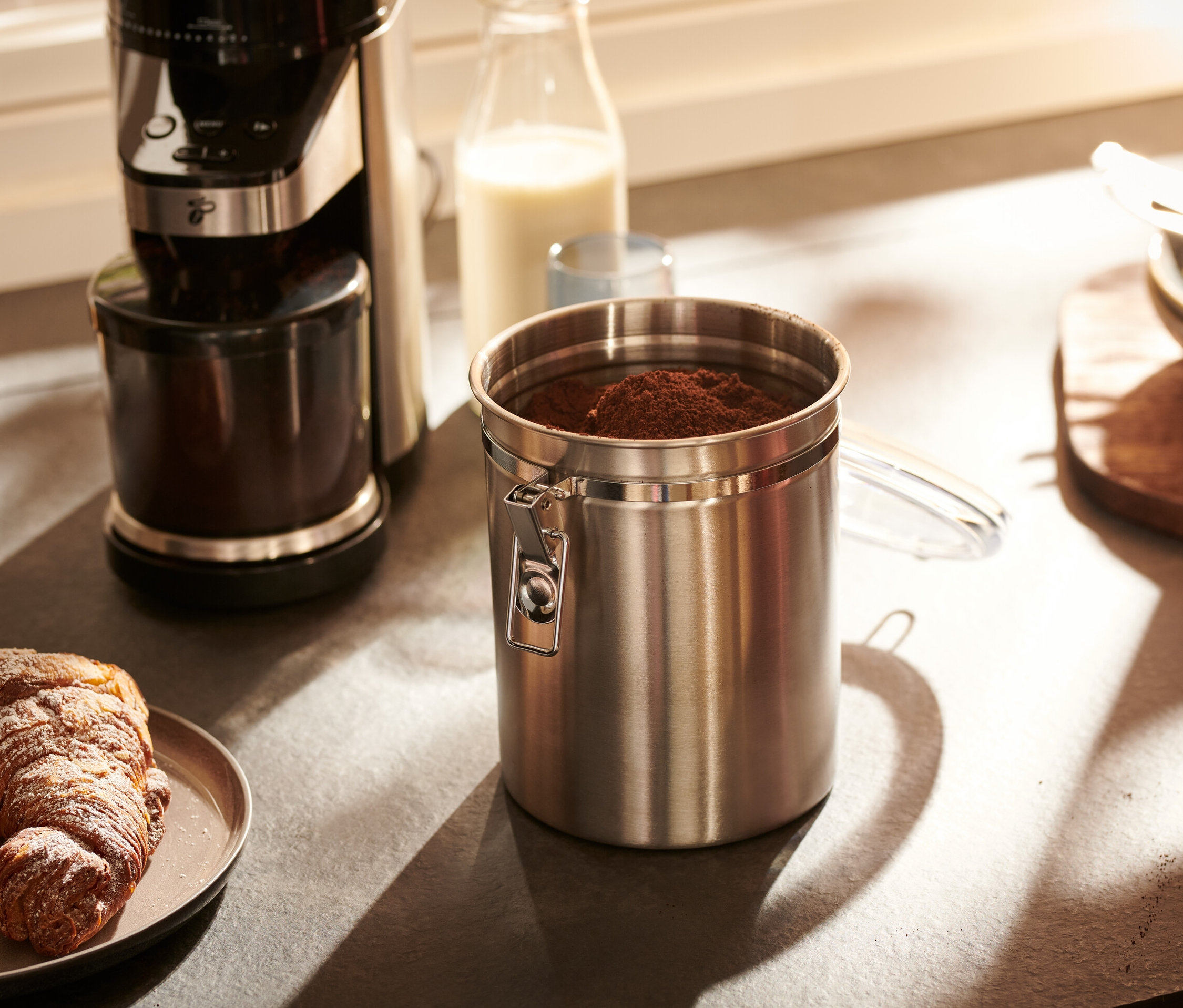 Eine Aromadose aus Edelstahl steht auf einer Küchentheke neben einer Kaffeemaschine und einem Teller mit einem Croissant.