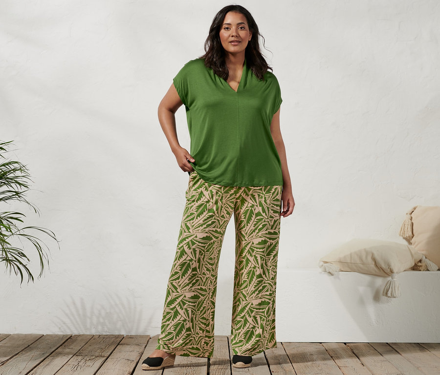 Eine Frau steht und trägt ein grünes Shirt mit Stehkragen, eine grün-beige bedruckte Palazzohose und Espadrilles mit Keilabsatz.