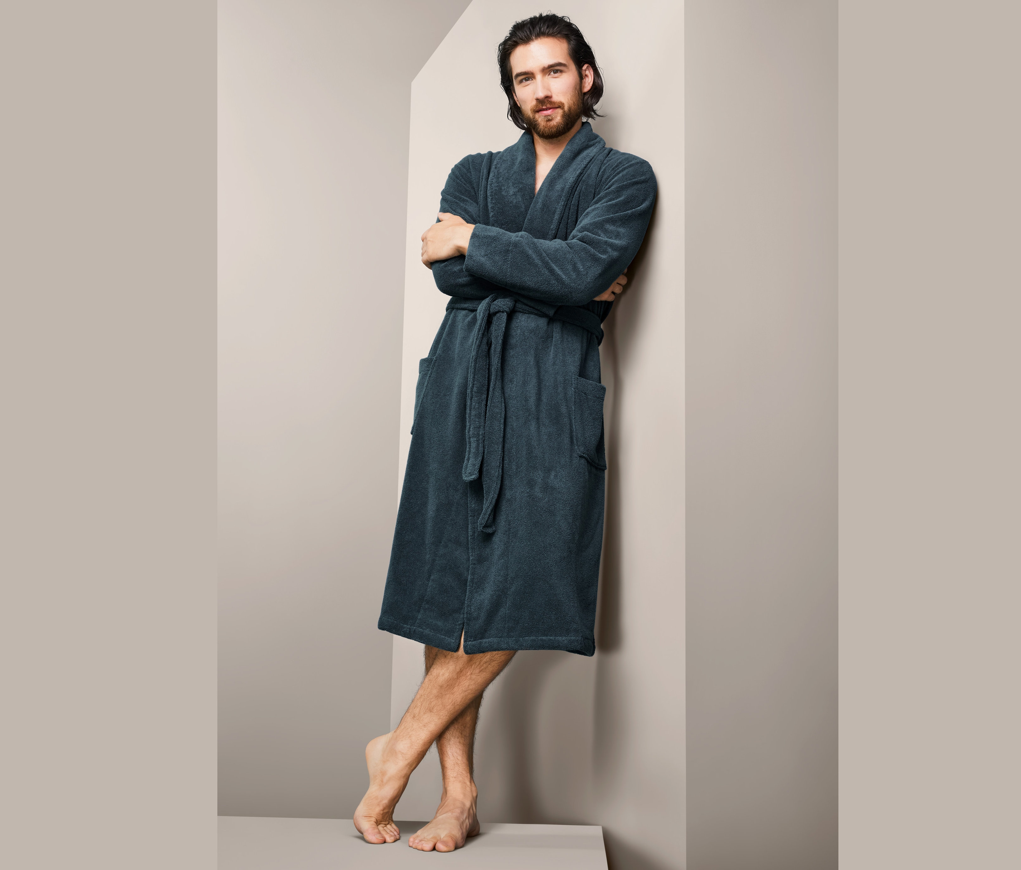 Mann mit braunen Haaren lehnt an einer Wand und trägt einen Herren-Bademantel.