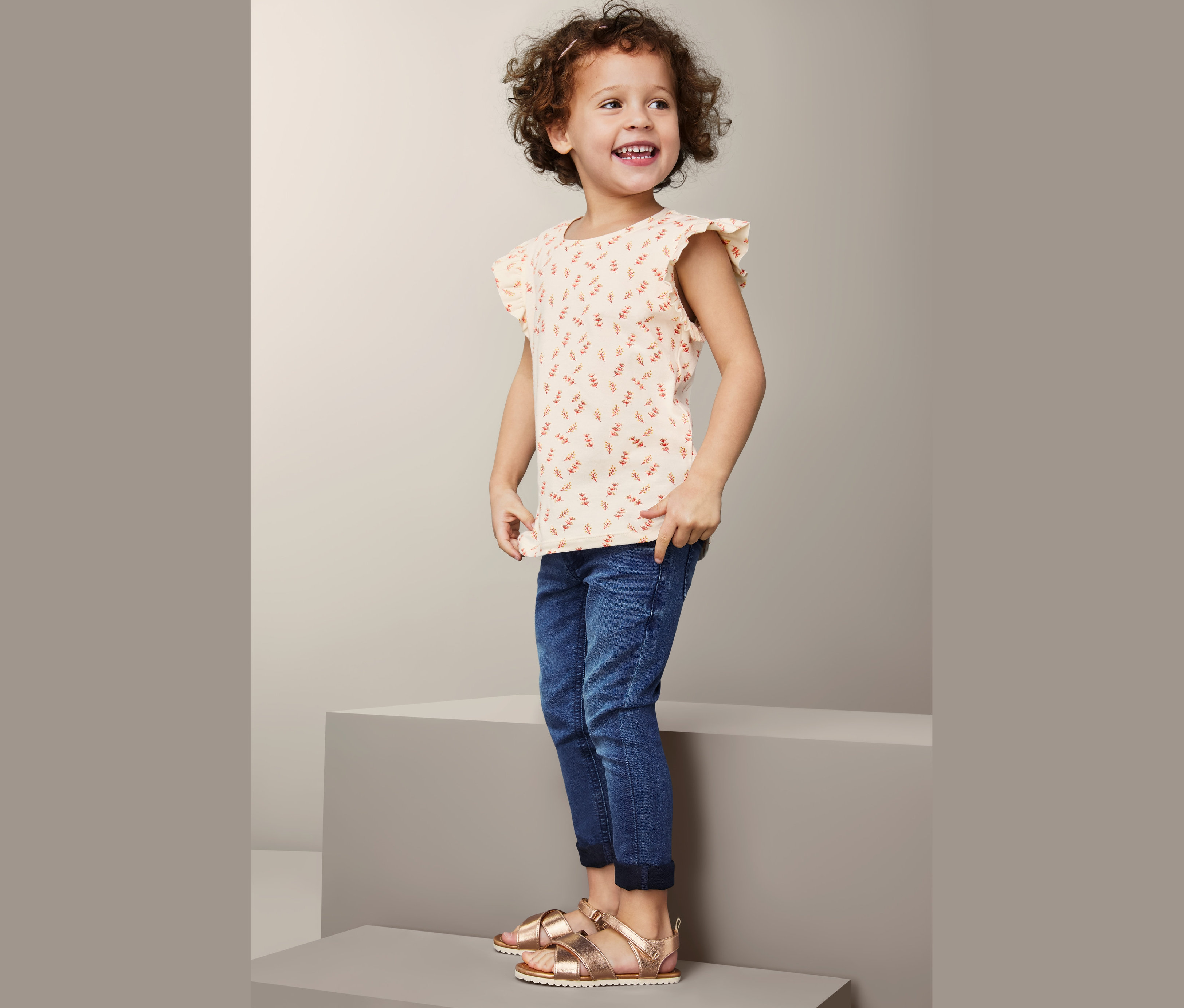 Ein lächelndes kleines Mädchen steht da und trägt ein gemustertes T-Shirt und Jeans. Sie trägt auch Sandalen.