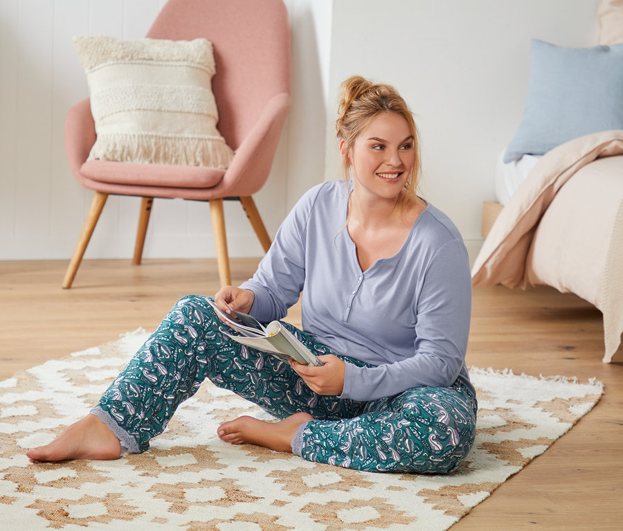 Frau sitzt auf dem Boden auf einem Teppich und liest ein Buch. Sie trägt einen Pyjama mit Alloverprint.