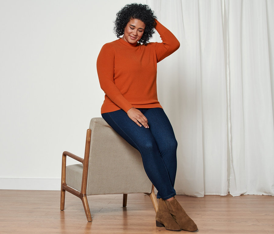 Frau sitzt auf einem Stuhl und trägt einen Strickpullover, Jeggings und Stiefelette aus Veloursleder.