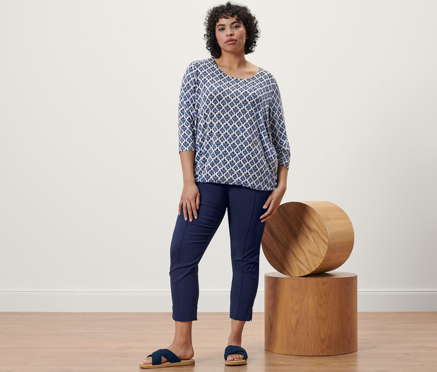 Frau steht und trägt ein Blusenshirt mit Alloverprint, eine Stretchhose in Dunkelblau und Komfortsandalen.