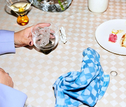 Auf einer karierten Tischdecke liegen ein Glas mit Eis, Essensreste auf einem Teller und eine blaue Serviette. Außerdem sind 4 Serviettenringe zu sehen.