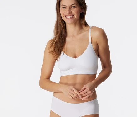 Lächelnde Frau mit braunen Haaren trägt ein weißes SCHIESSER Invisible Bustier mit Pads und weiße Slips, Hände gefaltet.