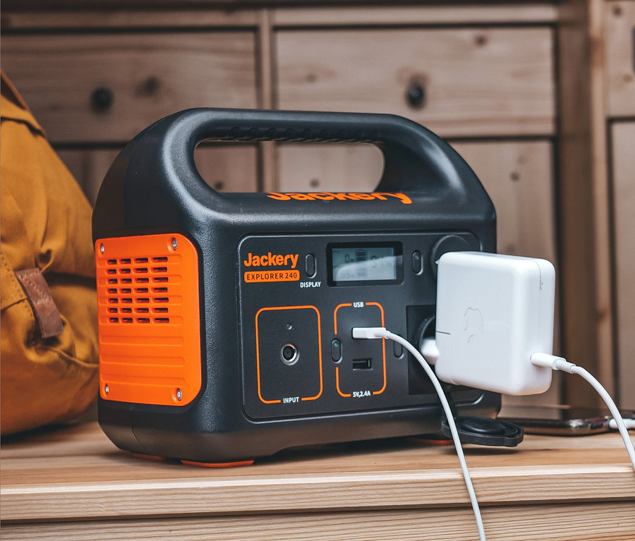 Auf einem Holzregal steht eine schwarz-orangefarbene Jackery Explorer 240 Ladestation mit Ladegerät und Kabel.