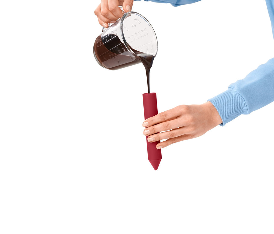 Frau giesst Schokolade aus einem Messbecher in einen roten Dekorierstift. Auf dem Boden liegt ein grüner Stift.