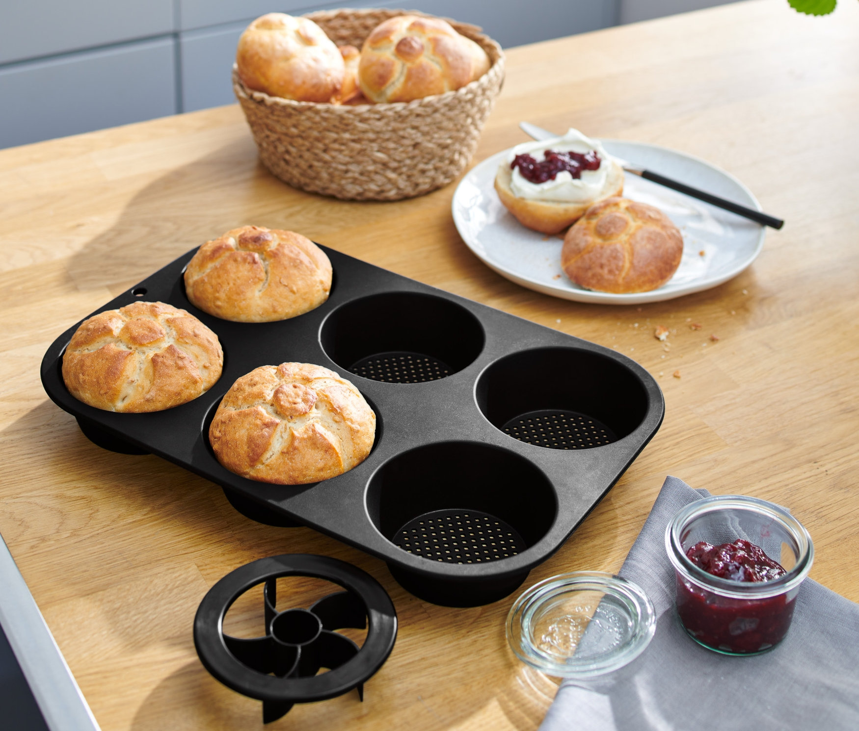 Vier Brötchen in einer schwarzen perforierten Brötchenbackform auf einem Holztisch.