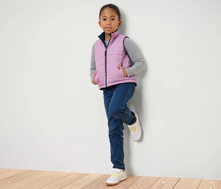 Mädchen, an eine Wand gelehnt, trägt eine Kinder-Wende-Steppweste, Jeans und Turnschuhe.