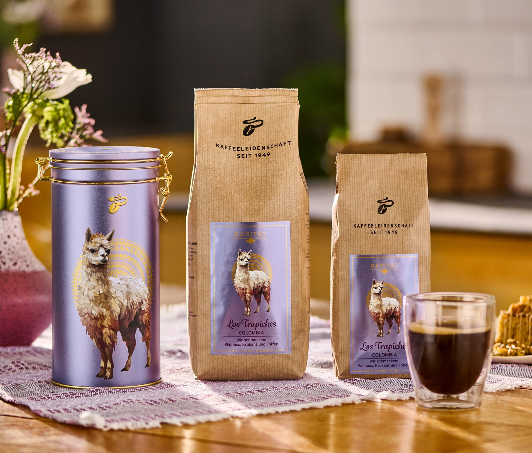 Auf einem Tisch stehen eine Blumenvase, eine Kaffeedose "Los Trapiches Kolumbien", zwei Kaffeepackungen, eine Tasse Kaffee und ein Teller mit Kuchen.