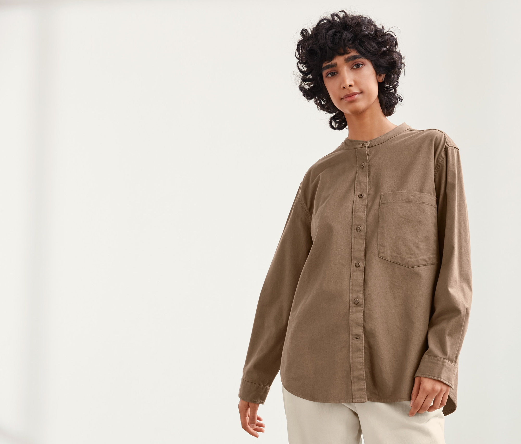 Frau posiert vor hellem Hintergrund in einer haselnussbraunen NAH/STUDIO Hemdbluse aus nachhaltiger Baumwolle und weißen Hosen.