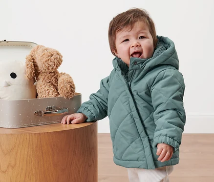 Kleinkind in Kleinkind-Steppjacke steht neben einem Koffer mit Spielzeug.