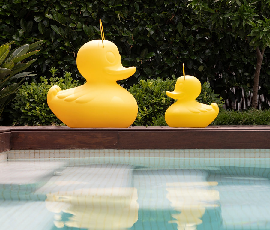 Zwei gelbe Goodnight Light LED-Leuchten »Ente« XL und S stehen auf einer Holzterrasse neben einem Pool.