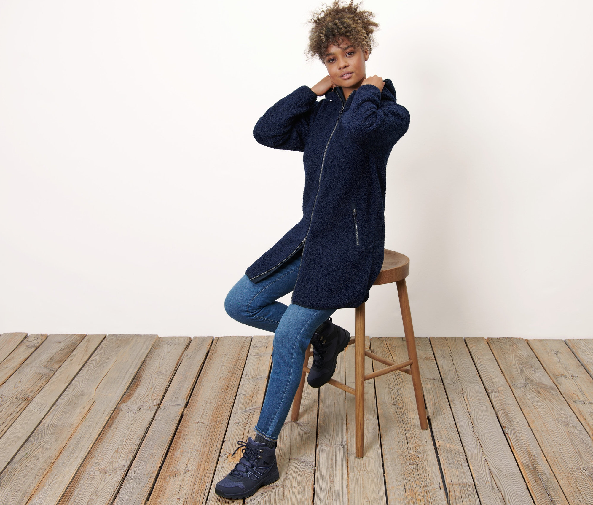 Frau sitzt auf einem Hocker und trägt einen Kapuzen-Teddy-Mantel.