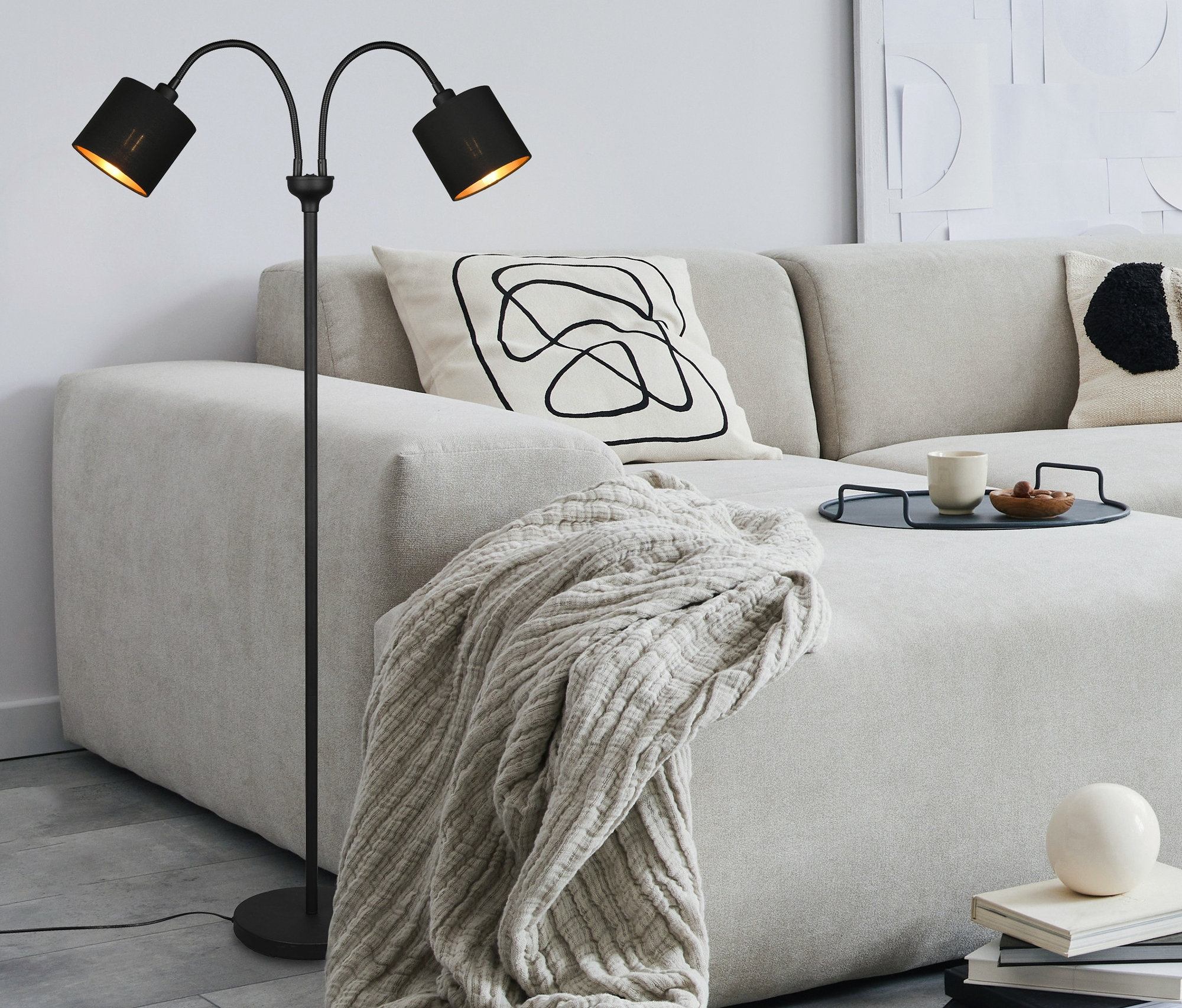Eine schwarze Reality Stehleuchte »Tommy« steht neben einem beigen Sofa mit Kissen und einer Decke.