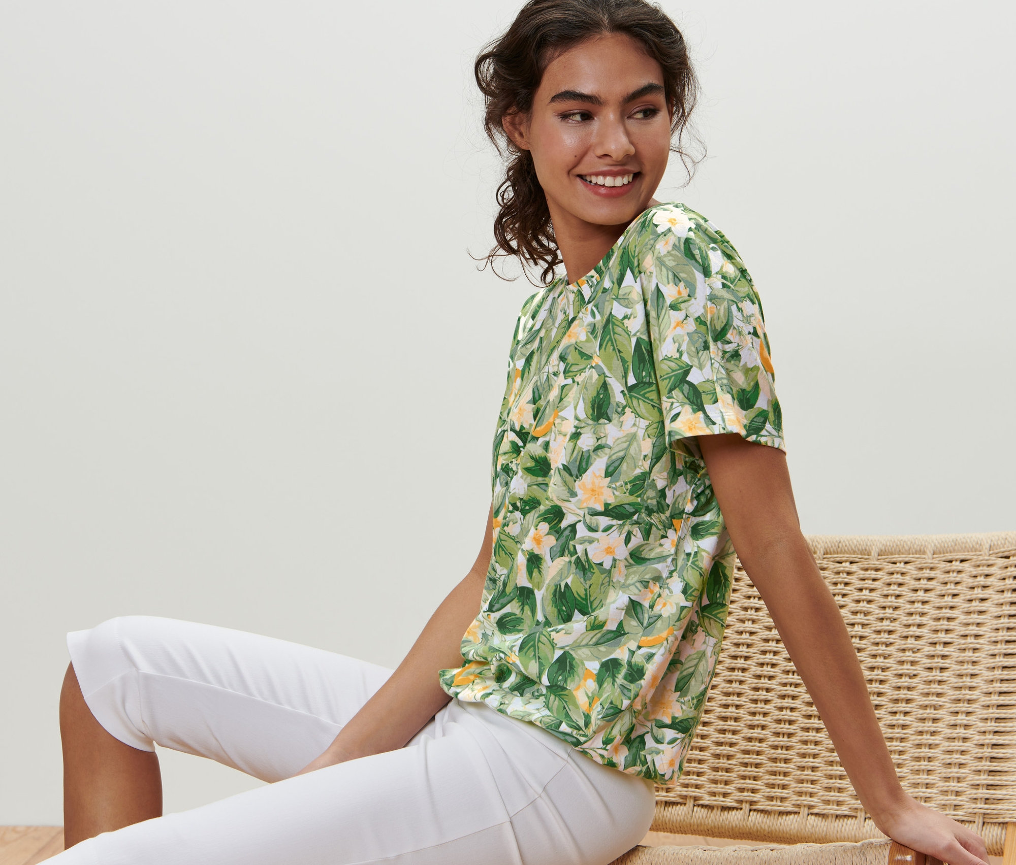Frau sitzt auf einem Korbstuhl und trägt ein bedrucktes Blusenshirt.