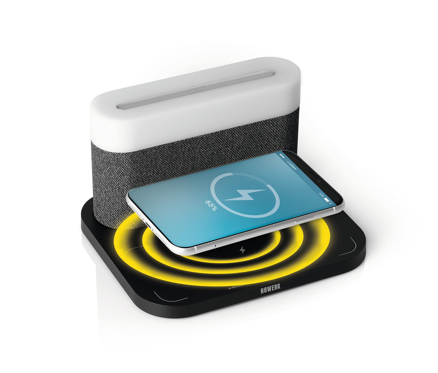 Ein Handy wird auf einem schwarzen N8WERK Wireless Charger neben einem grau-weißen Nachttischlampen-Wecker von N8WERK aufgeladen.
