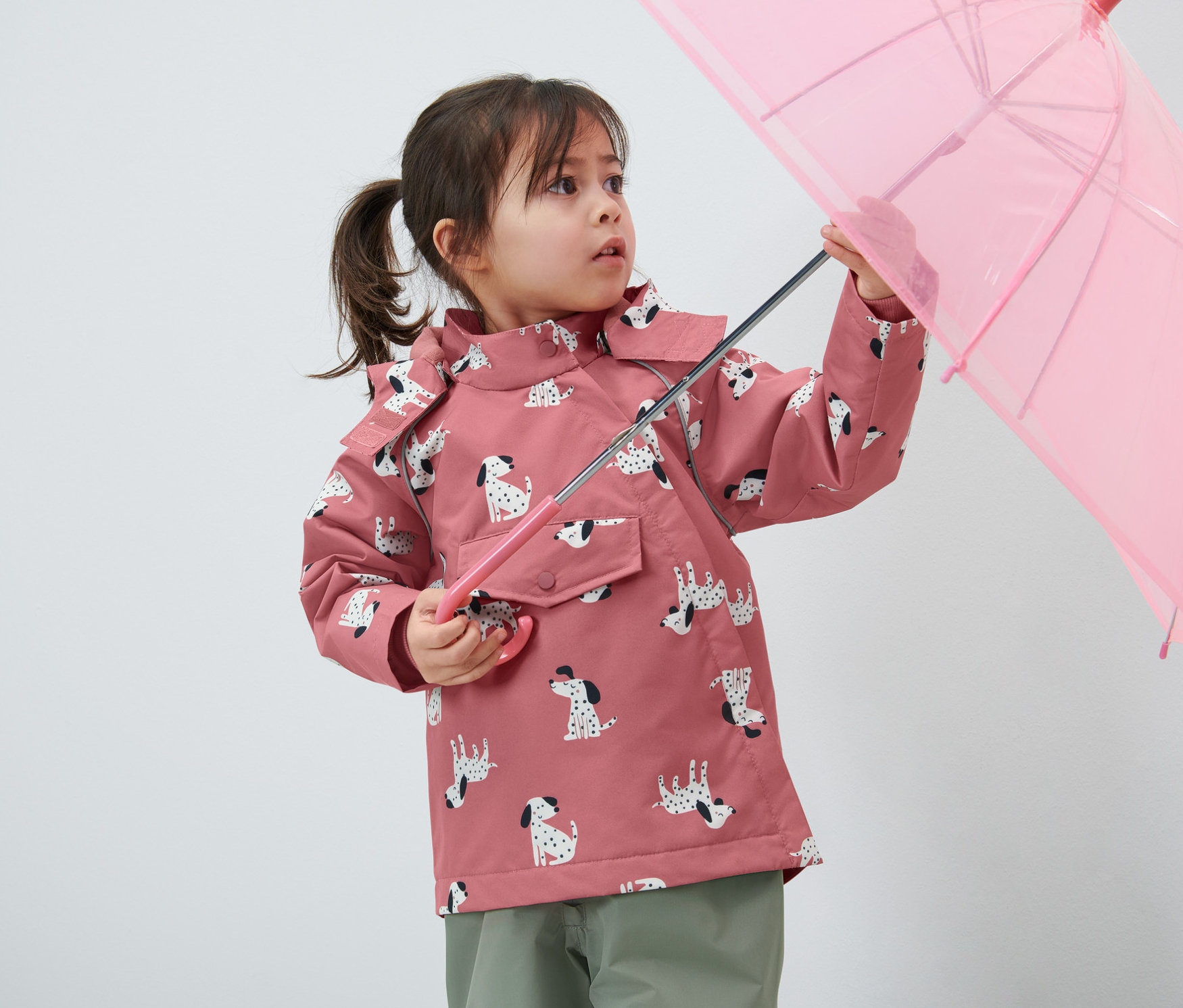 Mädchen hält einen rosa Regenschirm und trägt eine Kinder-Thermo-Regenjacke mit Hundemuster.