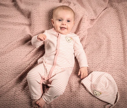 Baby liegt auf rosa Strickdecke und trägt einen rosa gestreiften Steiff Baby-Strampler mit Knopfleiste.