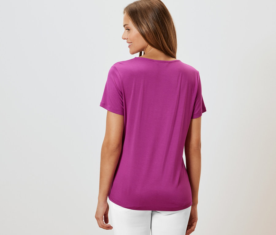 Frau von hinten in einem Shirt mit Faltendetail, berry und weißen Hosen.