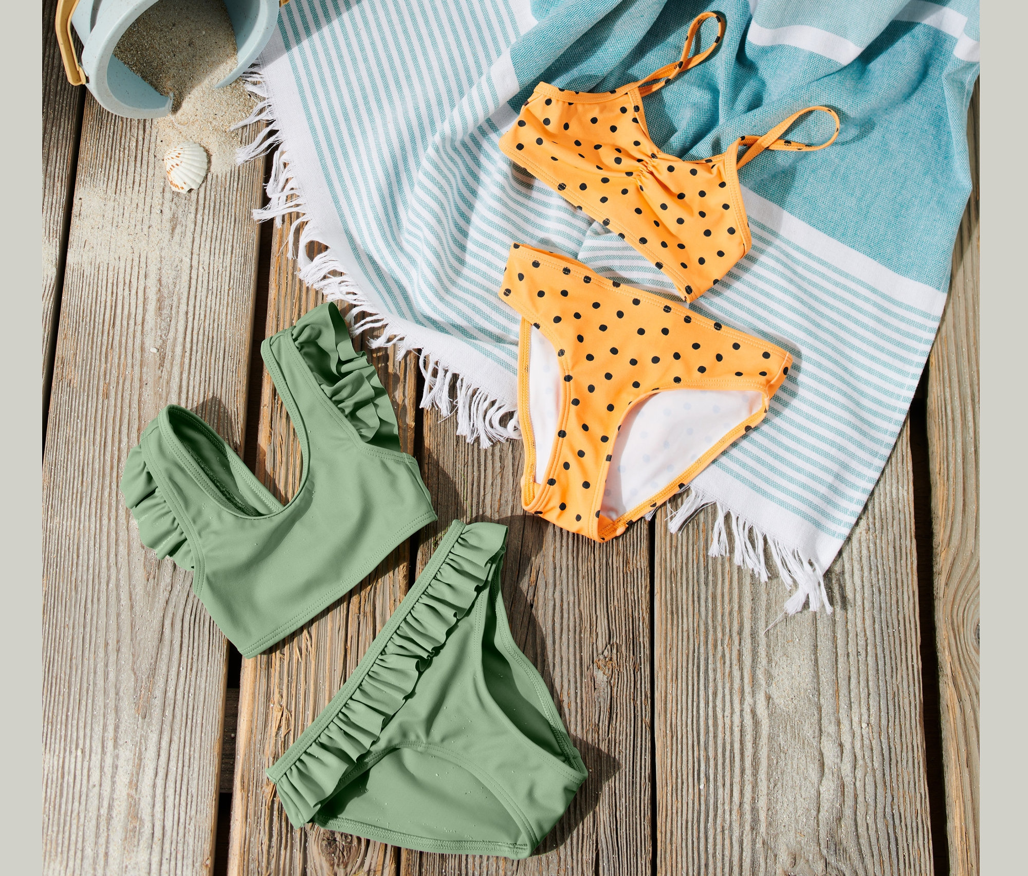 Auf einem Holzhintergrund liegen Kinderschwimmanzüge in zwei Ausführungen, orange mit schwarzen Punkten und grün. Ebenfalls zu sehen sind ein Strandtuch mit blauen Streifen und ein Eimer mit Sand.