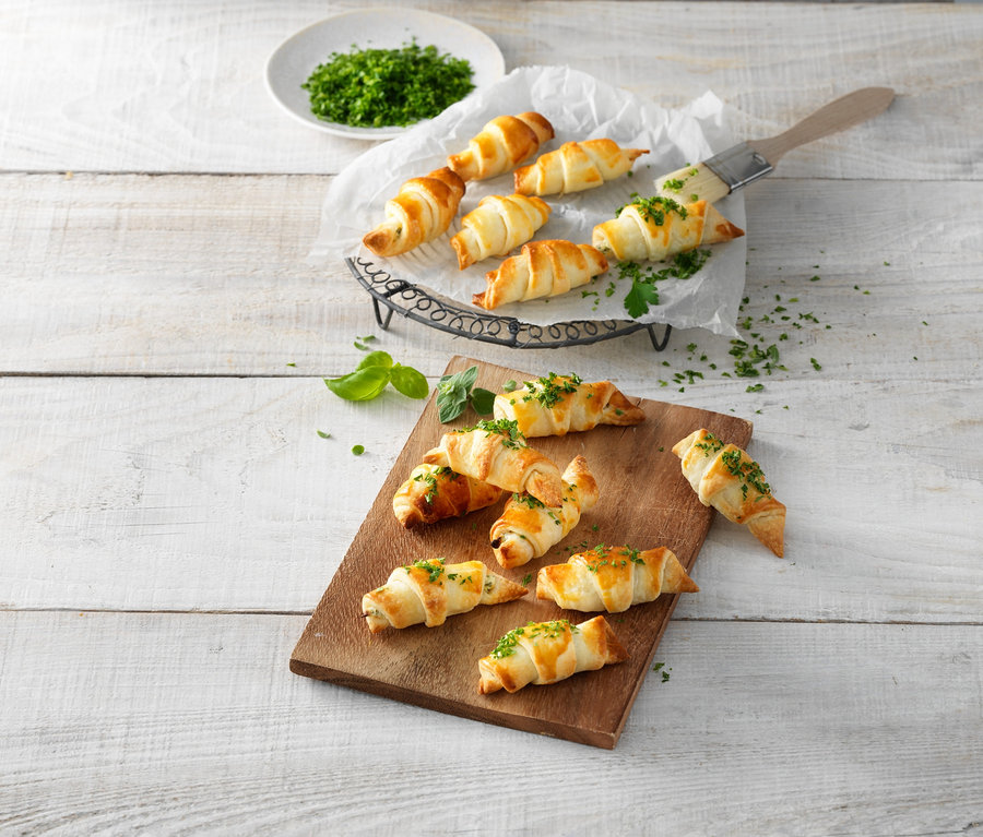 Mit Kräutern bestreute Croissants auf einem Holzbrett und einem Drahtständer. Croissant-Roller.