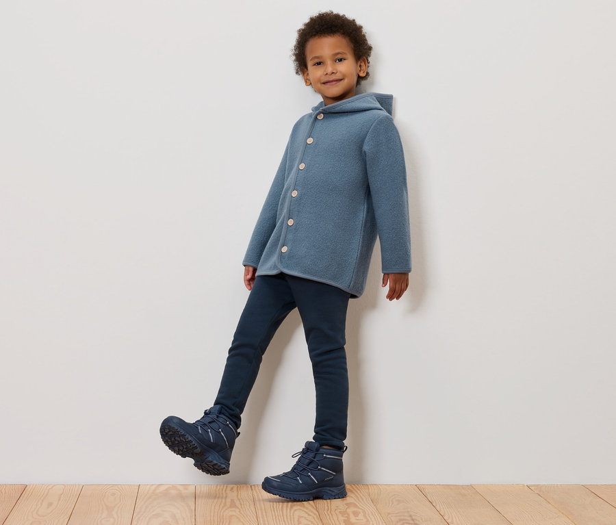 Ein Junge in einem blauen Wollmantel mit Kapuze, dunkelblauen Jeans und blauen Winterstiefeln posiert an einer Wand.