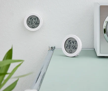 Zwei weiße Digitaluhren. Eine Uhr hängt an der Wand. Die zweite Uhr steht auf einem Tisch neben einem weißen Radio.