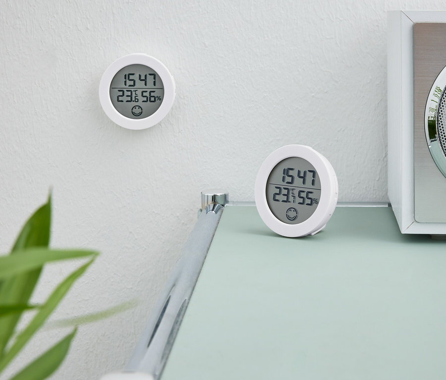 Zwei weiße Digitaluhren. Eine Uhr hängt an der Wand. Die zweite Uhr steht auf einem Tisch neben einem weißen Radio.