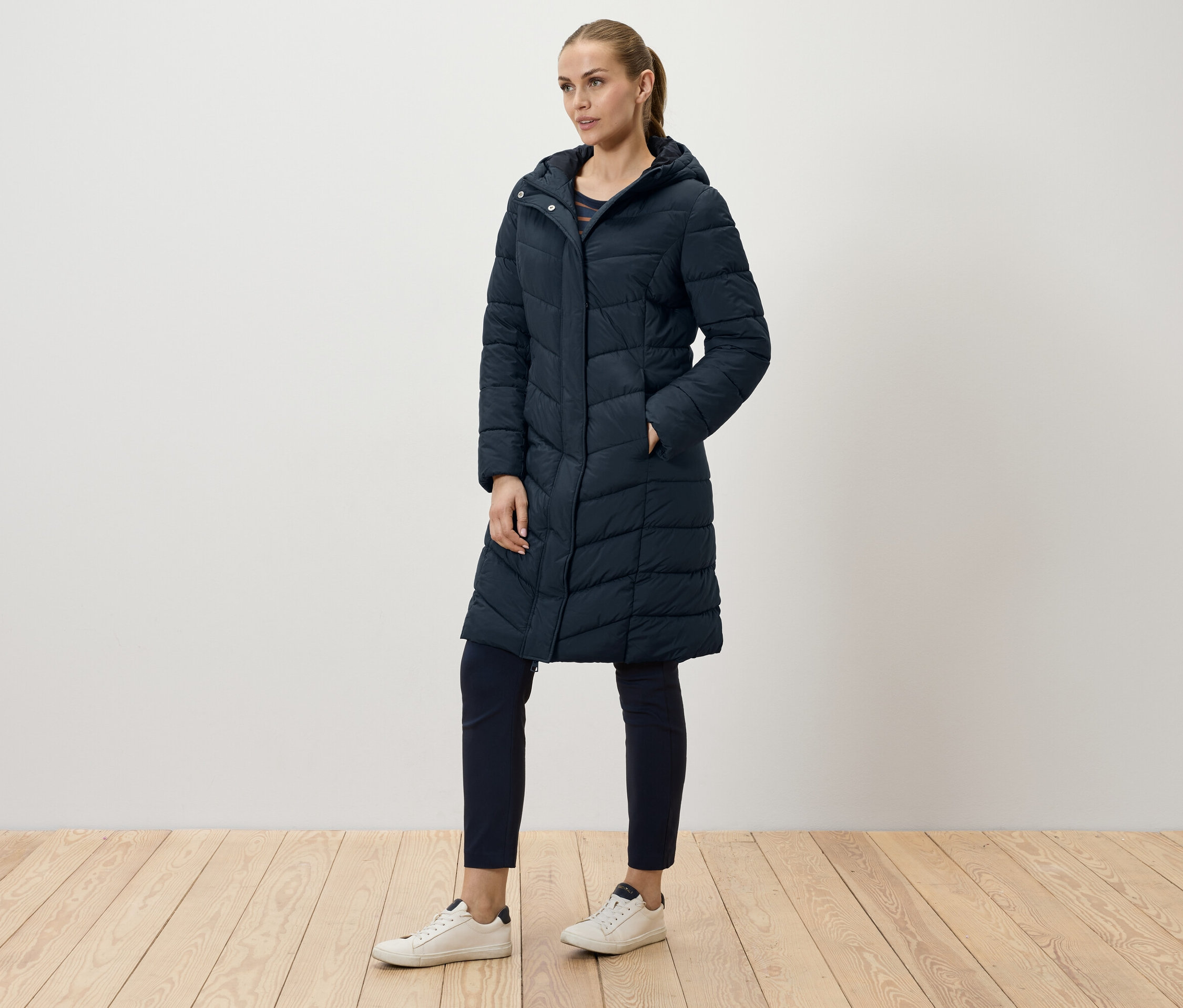 Eine Frau posiert in einem langen, marineblauen Steppmantel, marineblauen Hosen und weißen Turnschuhen.