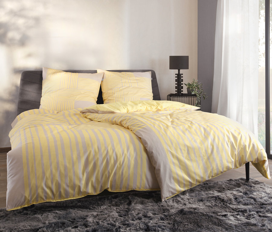 Gelb-grau gestreifte SCHÖNER WOHNEN-Kollektion Bettwäsche auf einem Bett mit grauem Kopfteil.