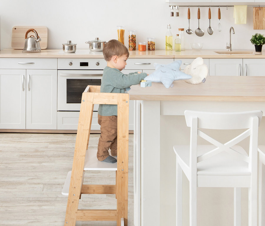 Ein Junge steht auf dem weißen roba Lernturm aus Holz in einer Küche und spielt mit Spielsachen.