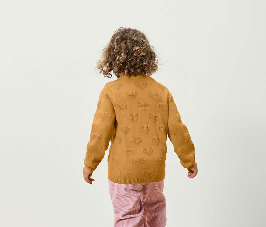 Ein Junge mit lockigem Haar steht mit dem Rücken zur Kamera und trägt einen gelben Pullover mit Herzmuster und eine rosafarbene Hose.