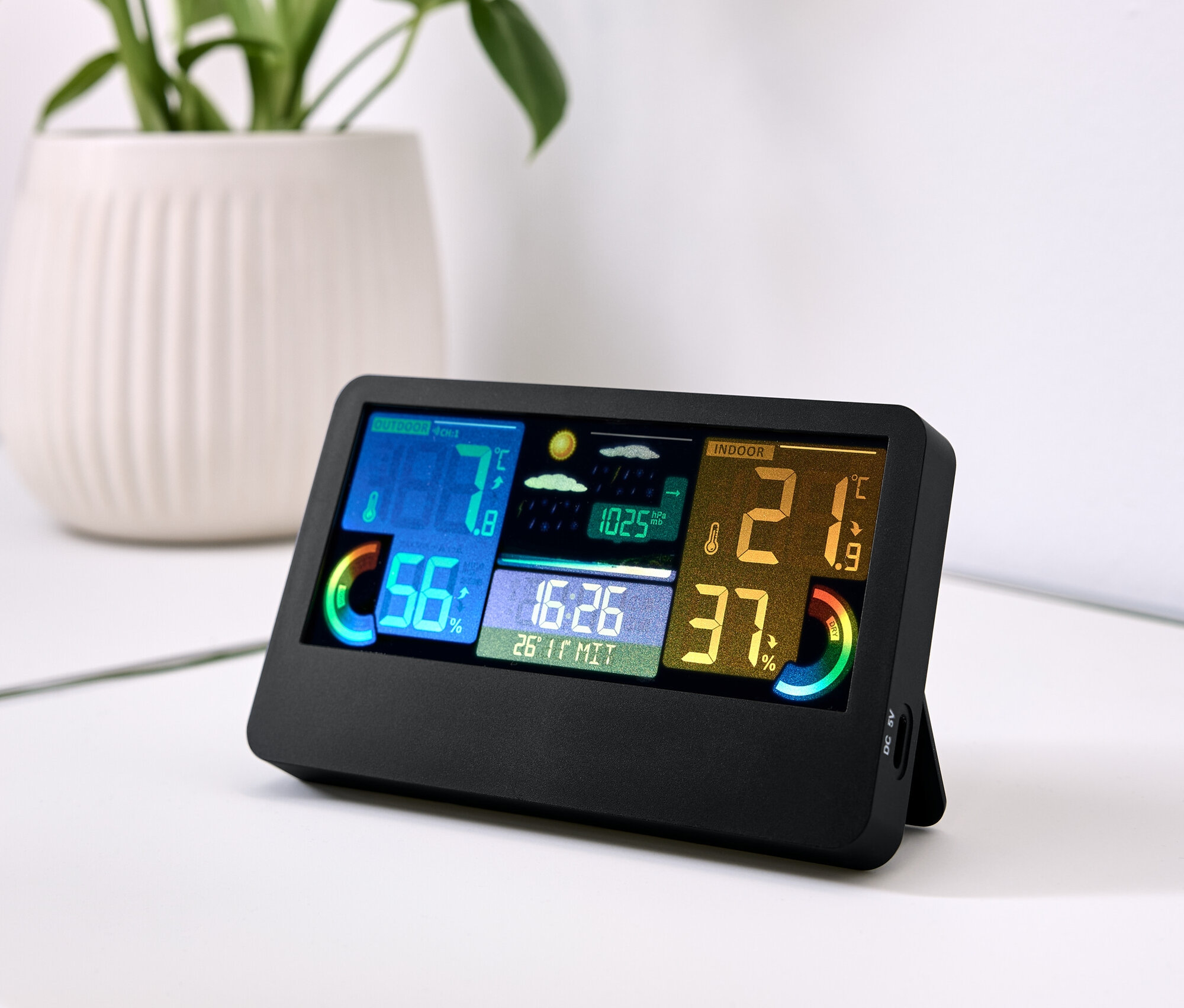 Eine schwarze Wetterstation mit farbigem Display zeigt Temperatur, Luftfeuchtigkeit und Zeit an. Daneben liegen Schlüssel und im Hintergrund eine Pflanze in einem weißen Topf.