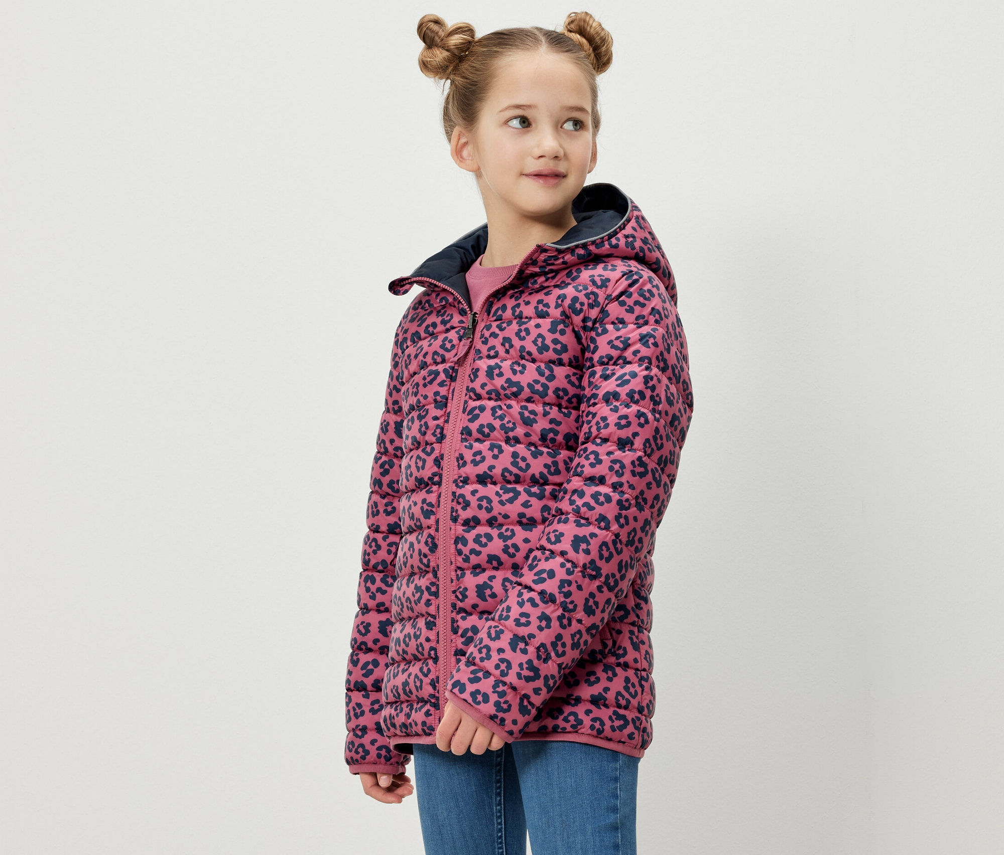 Mädchen posiert in einer 2-in-1-Kinder-Softshell- und Steppjacke. Die Jacke hat einen rosafarbenen Hintergrund mit einem marineblauen Tiermuster.