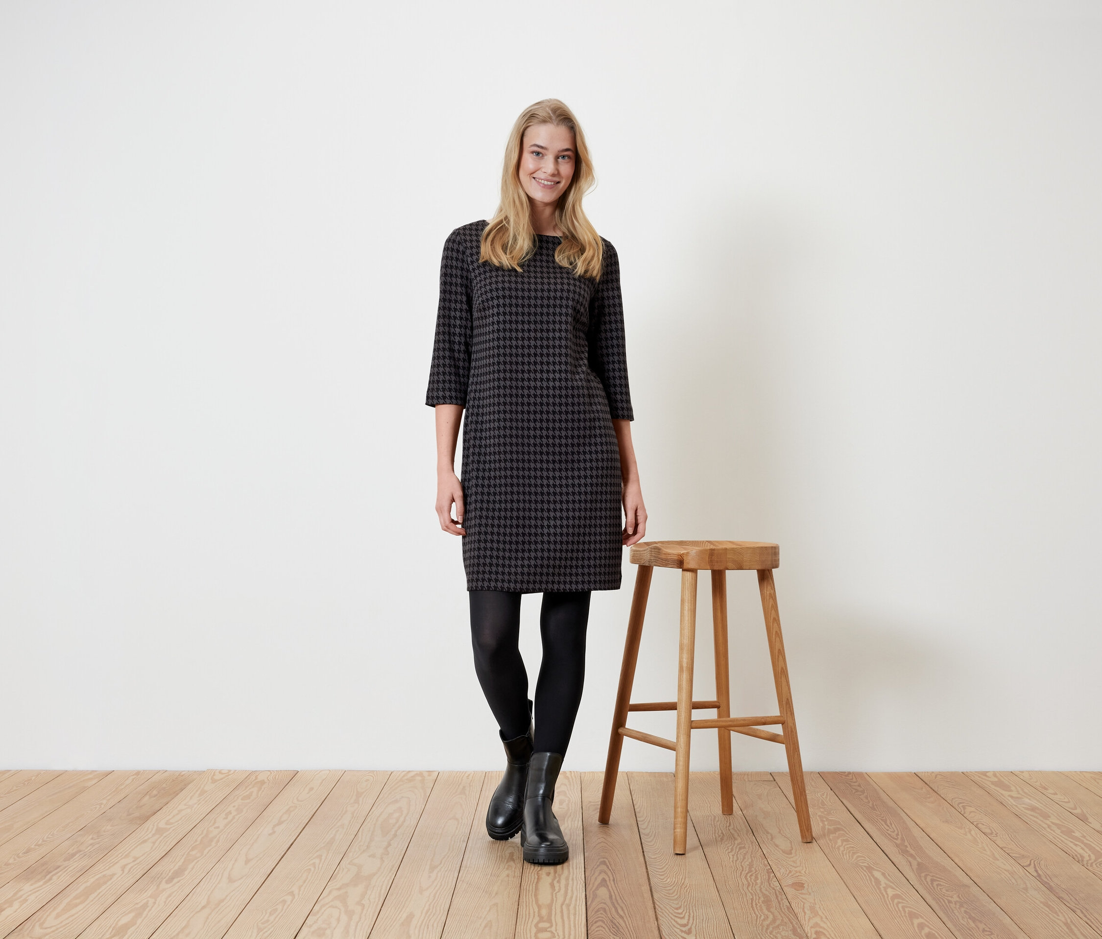 Frau steht neben einem Holzhocker und trägt ein Hahnentrittkleid, schwarze Leggings und Stiefel.