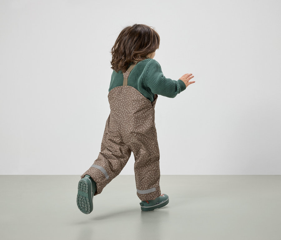 Ein Kind in braunen Latzhosen und grünem Pullover rennt vom Betrachter weg.
