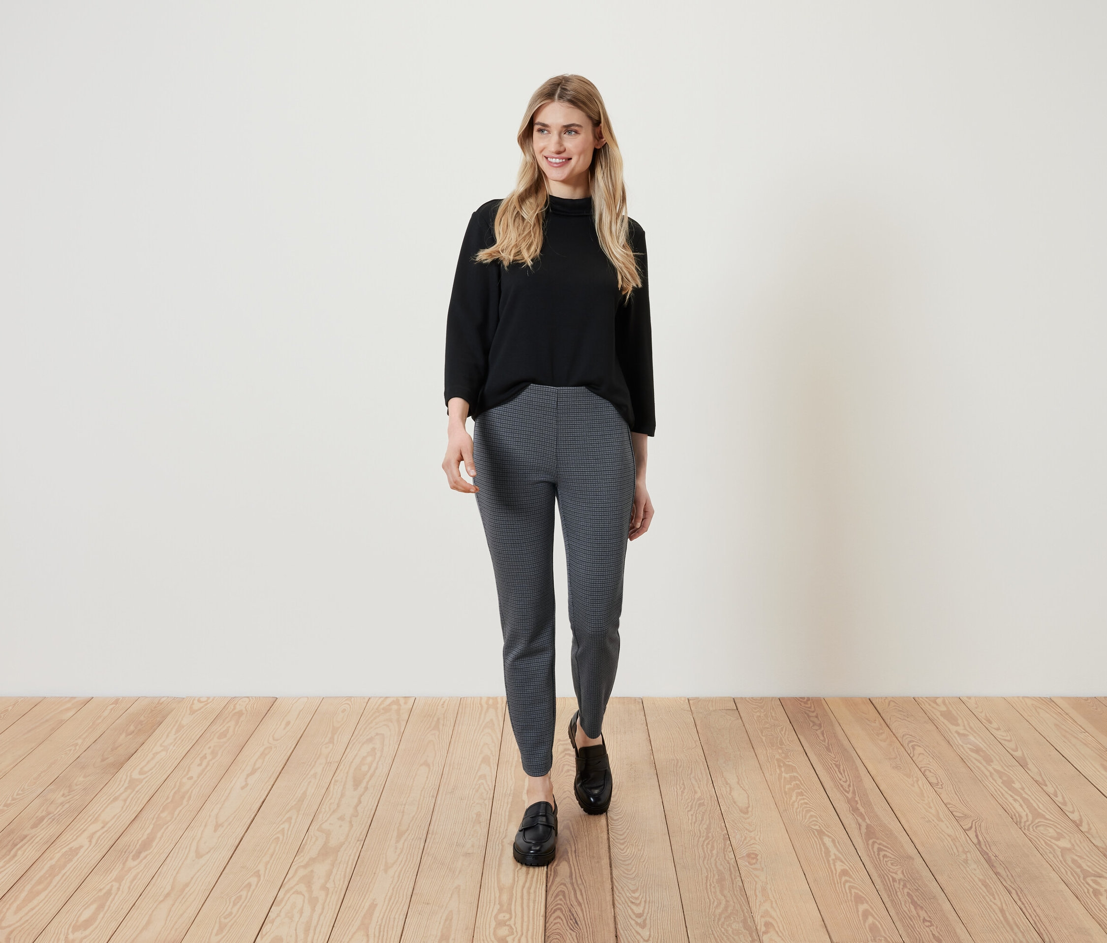 Eine blonde Frau in einem schwarzen Oberteil und gemusterten Hosen geht auf einem Holzboden.