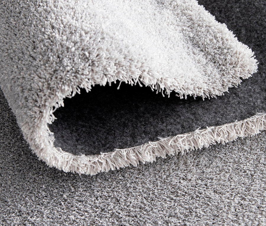 Nahaufnahme eines hellgrauen, zotteligen Teppichs »Mellow«. Ein Teil des Teppichs ist aufgerollt und gibt den Blick auf die dunkelgraue Unterseite und die Fransen frei.