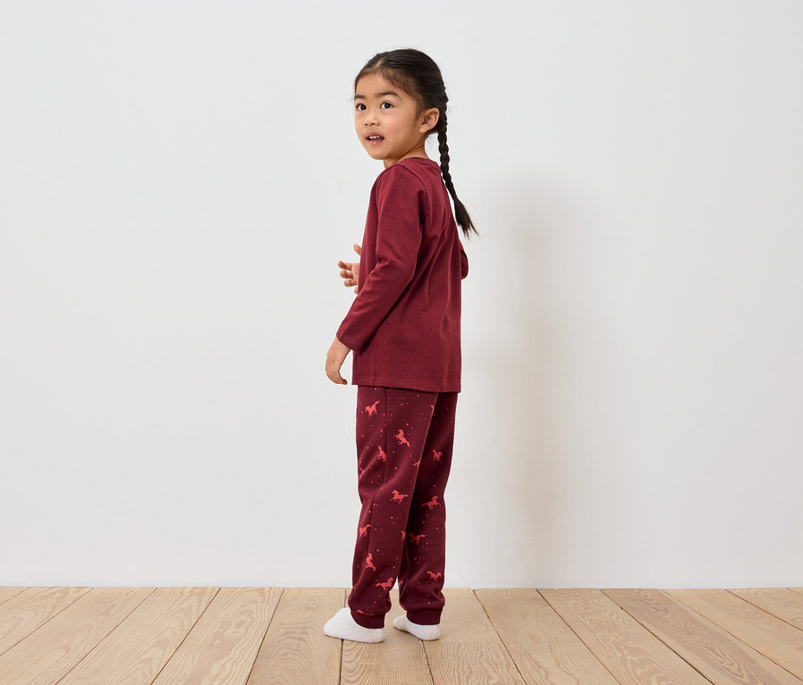 Ein asiatisches Mädchen mit Zopf steht seitlich und schaut über die Schulter zurück. Sie trägt einen bordeauxfarbenen Pyjama mit Pferdemotiv.