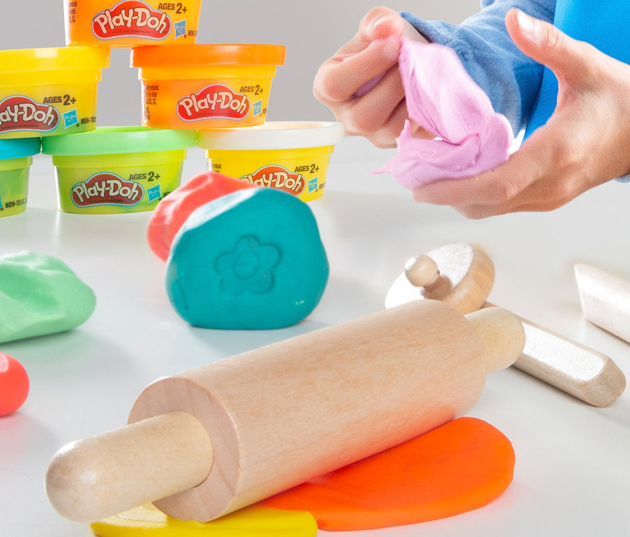 Kinderhände kneten rosa Play-Doh Knete. Im Hintergrund sind Becher mit Knete und eine Teigrolle aus Holz zu sehen. roba x Play-Doh Eisstand
