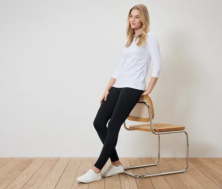 Blonde Frau sitzt seitlich auf einem Stuhl. Sie trägt ein weißes Dreiviertelarm-Shirt, schwarze Leggings und weiße Turnschuhe.
