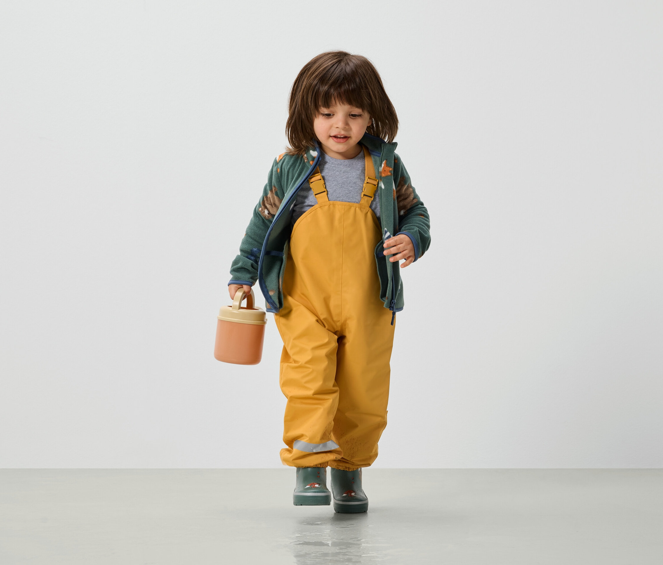 Kleinkind in gelber Latzhose, grüner Jacke und Gummistiefeln hält eine orangefarbene Lunchbox in der Hand.