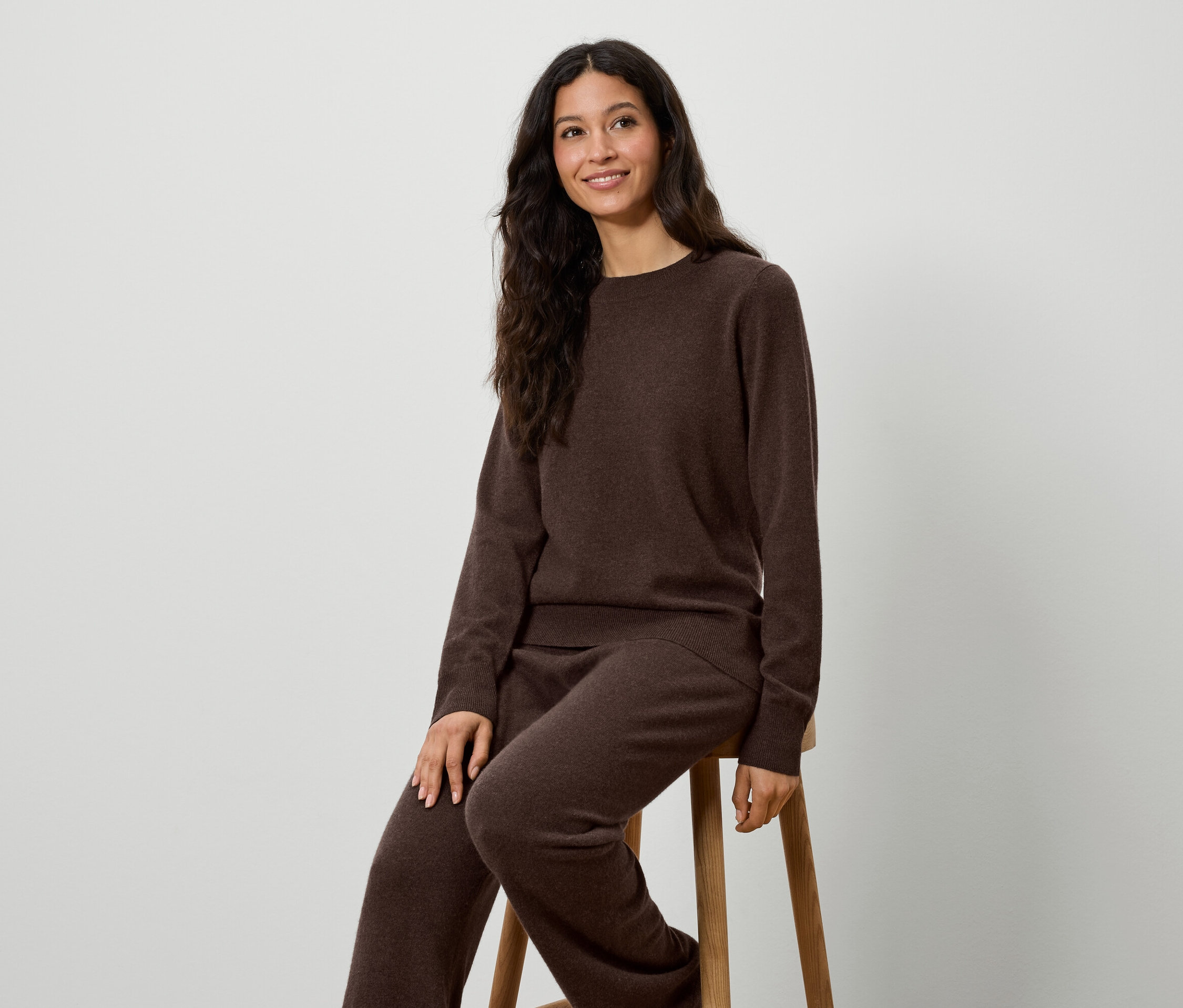 Eine Frau sitzt auf einem Hocker und trägt einen braunen Strickpullover und eine Hose.